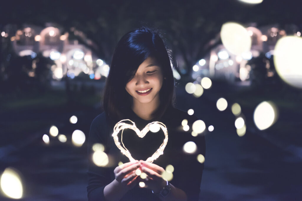 Young woman holding a heart-shaped string of lights at night, surrounded by bokeh.