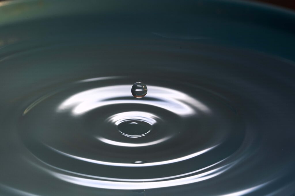 A single water droplet suspended above ripples on a dark, reflective surface.